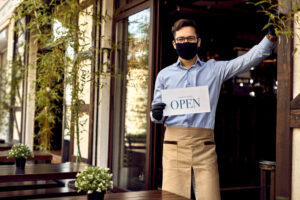 male server with glasses and brown apron and black face mask holding an open for business sign. he is standing on the patio of a cafe with brown chairs and tables.