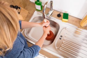 Woman Using Plunger In Blocked Kitchen Sink To Unclog Drain
