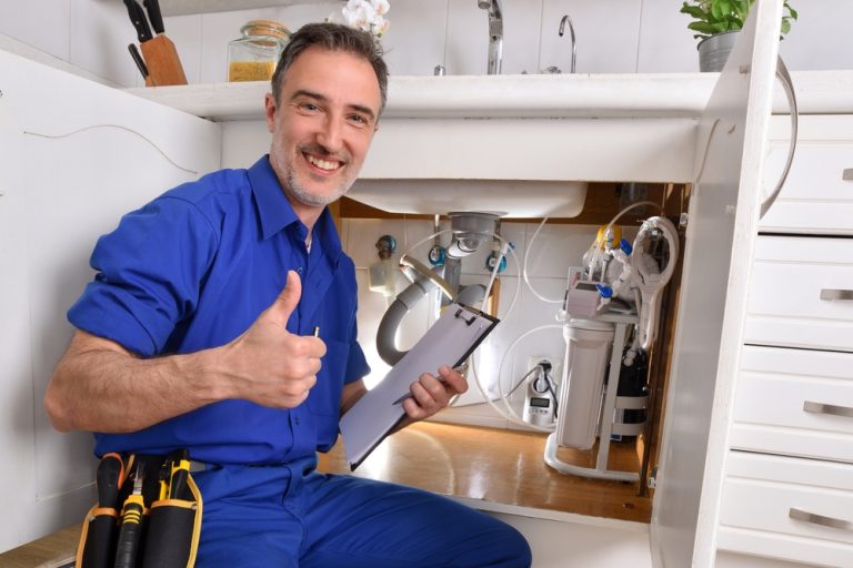 Plumbing technician checking plumbing under the sink of a home kitchen with notepad and hand with ok gesture.