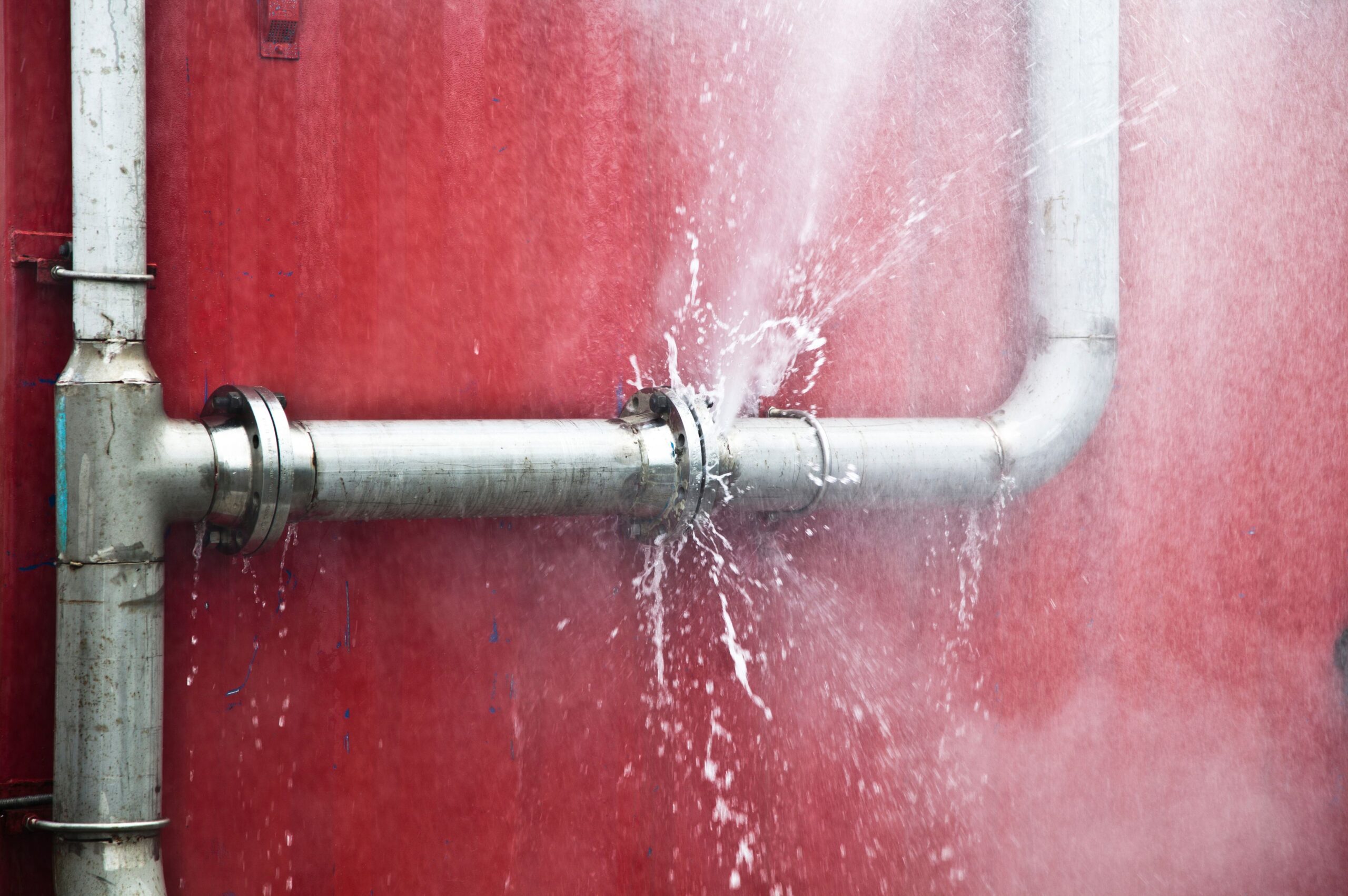 Water spraying from a burst metal water pipe against a red wall, indicating a major pipe leak.