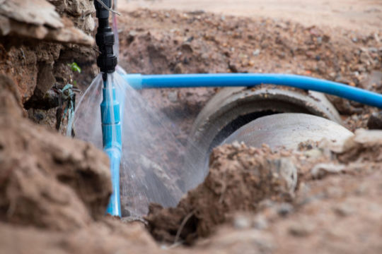 Underground blue water pipe leaking near a concrete drain, showing a water main break in progress.