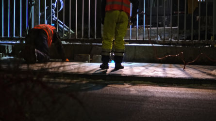 Workers performing trenchless sewer repair at night under artificial lighting on a city sidewalk.