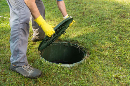 Technician lifting septic tank access lid for inspection and maintenance on a residential lawn.