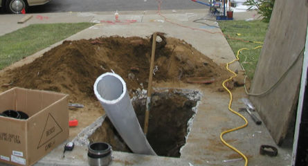 Construction site showing a small access hole being prepared for trenchless sewer line repair with a new pipe being inserted.